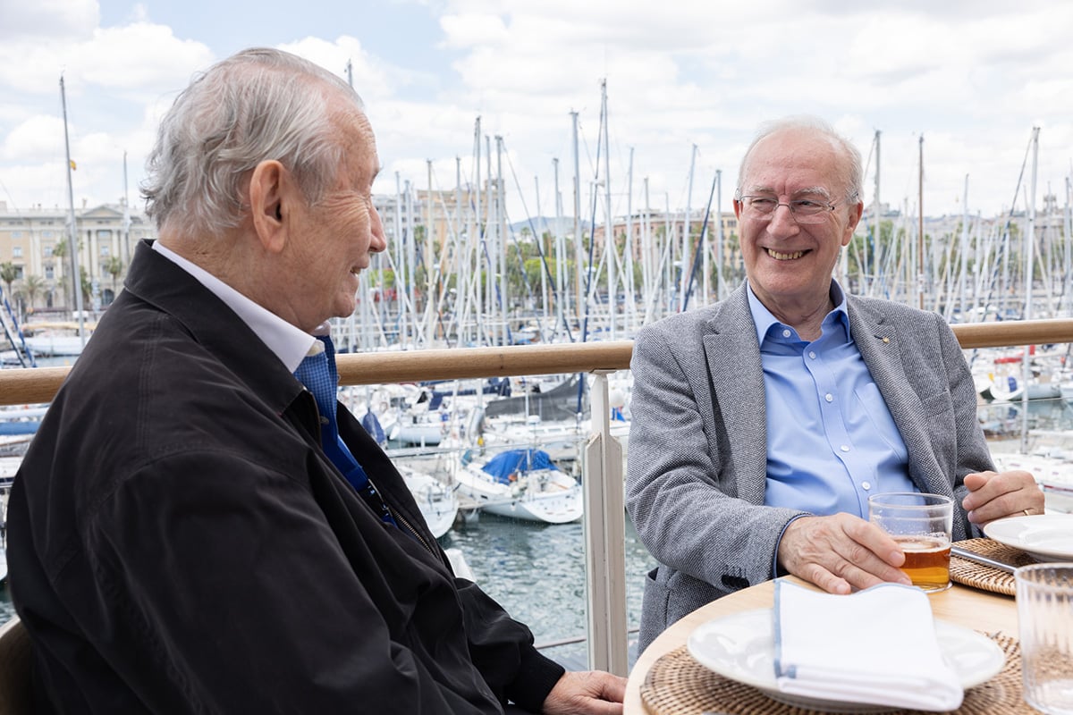 Two men sit at a table outdoors in front of many boat masts.