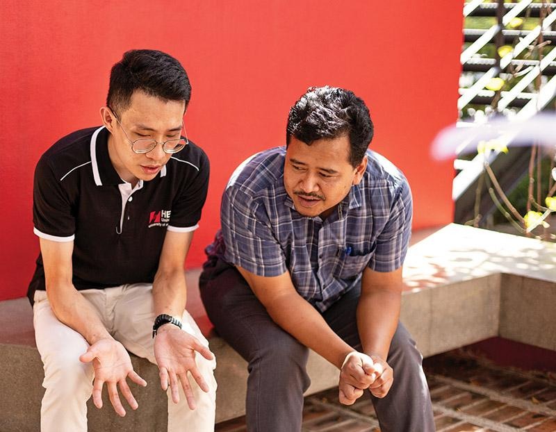Two people seated outdoors in conversation beside a red wall.