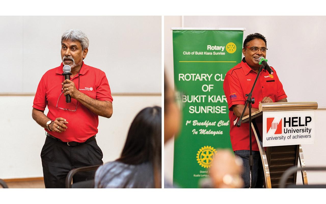 Person speaking into a microphone beside a lectern and Rotary event banner during a presentation.