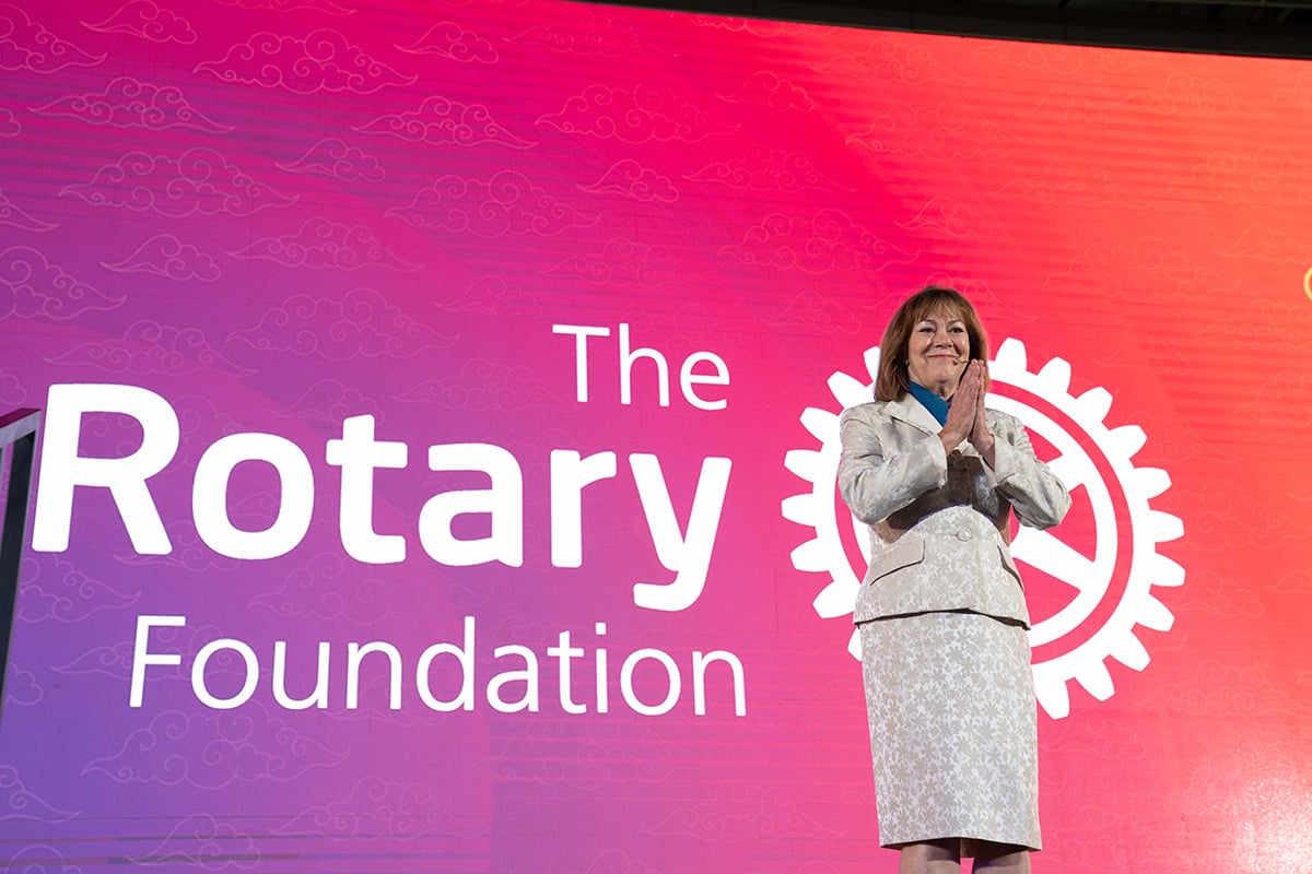 A woman in a gray suit stands in front of a large illuminated screen with The Rotary Foundation logo.
