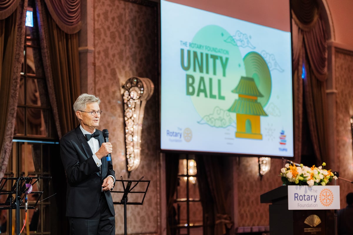 A man in a tuxedo speaks into a microphone near a screen saying The Rotary Foundation Unity Ball.