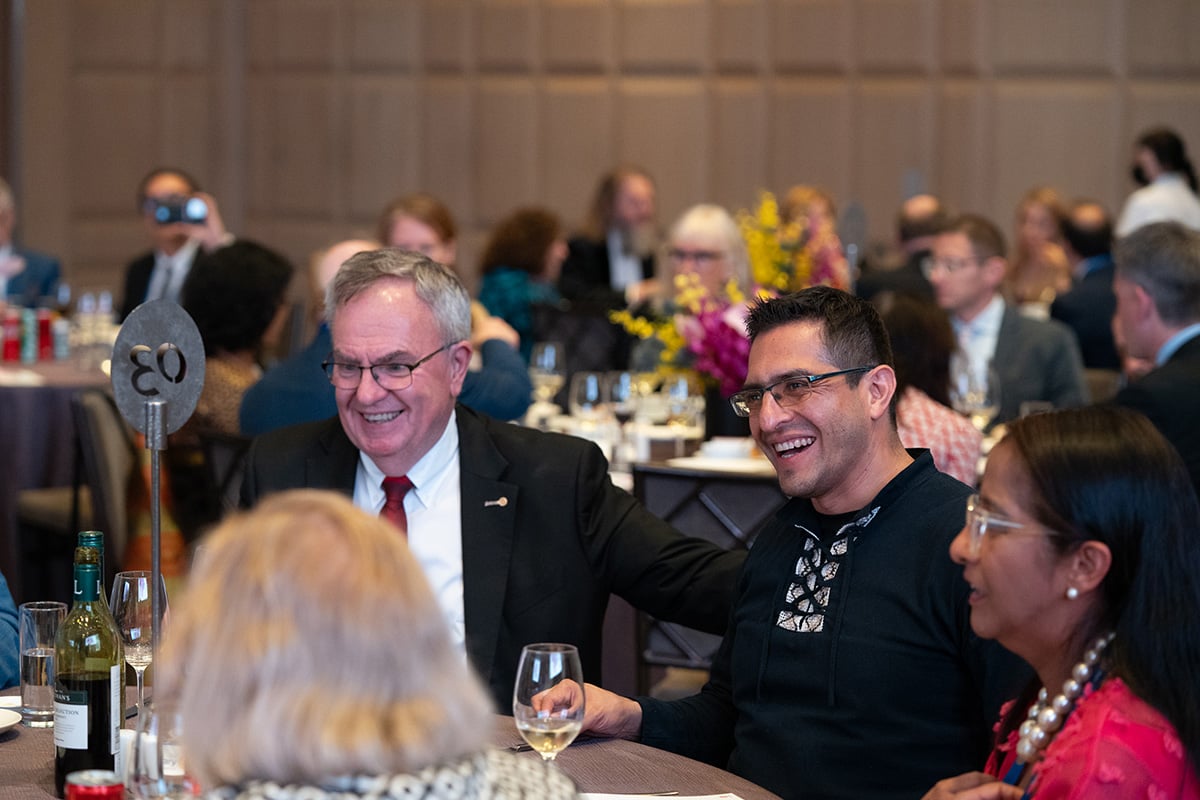 People seated at a table talk together. In the background, more people are seated at tables.