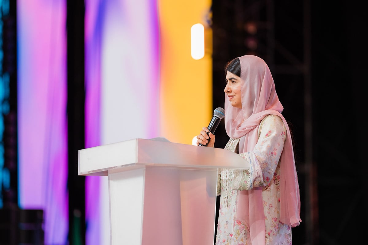 Malala Yousafzai, die jüngste Friedensnobelpreisträgerin aller Zeiten, spricht an einem Rednerpult, während sie ein Mikrofon in der Hand hält, auf einer Bühne mit farbenfrohen, beleuchteten Paneelen.