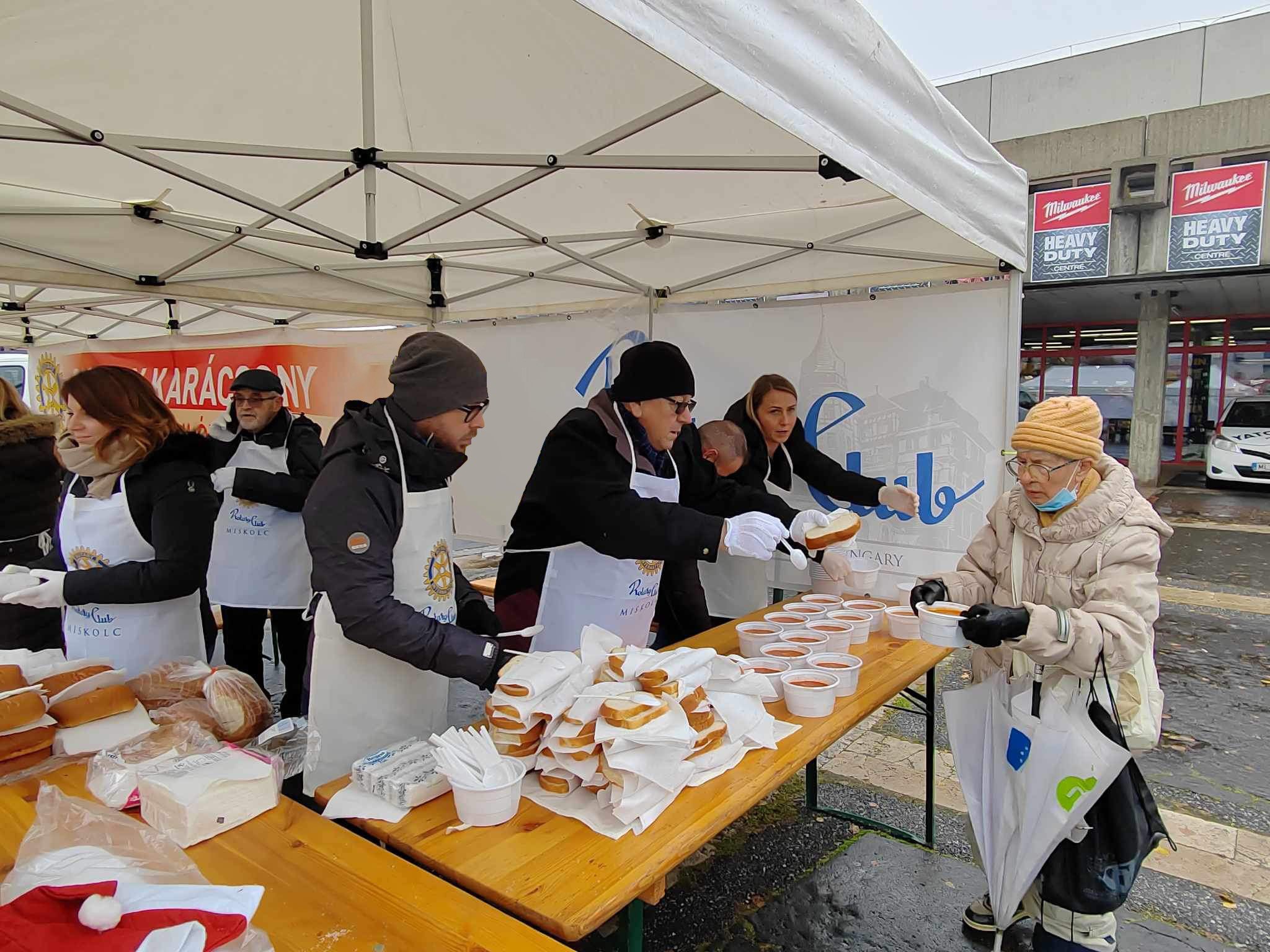 Volunteers distributing boxed meals and beverages from tables under a canopy at an outdoor event.