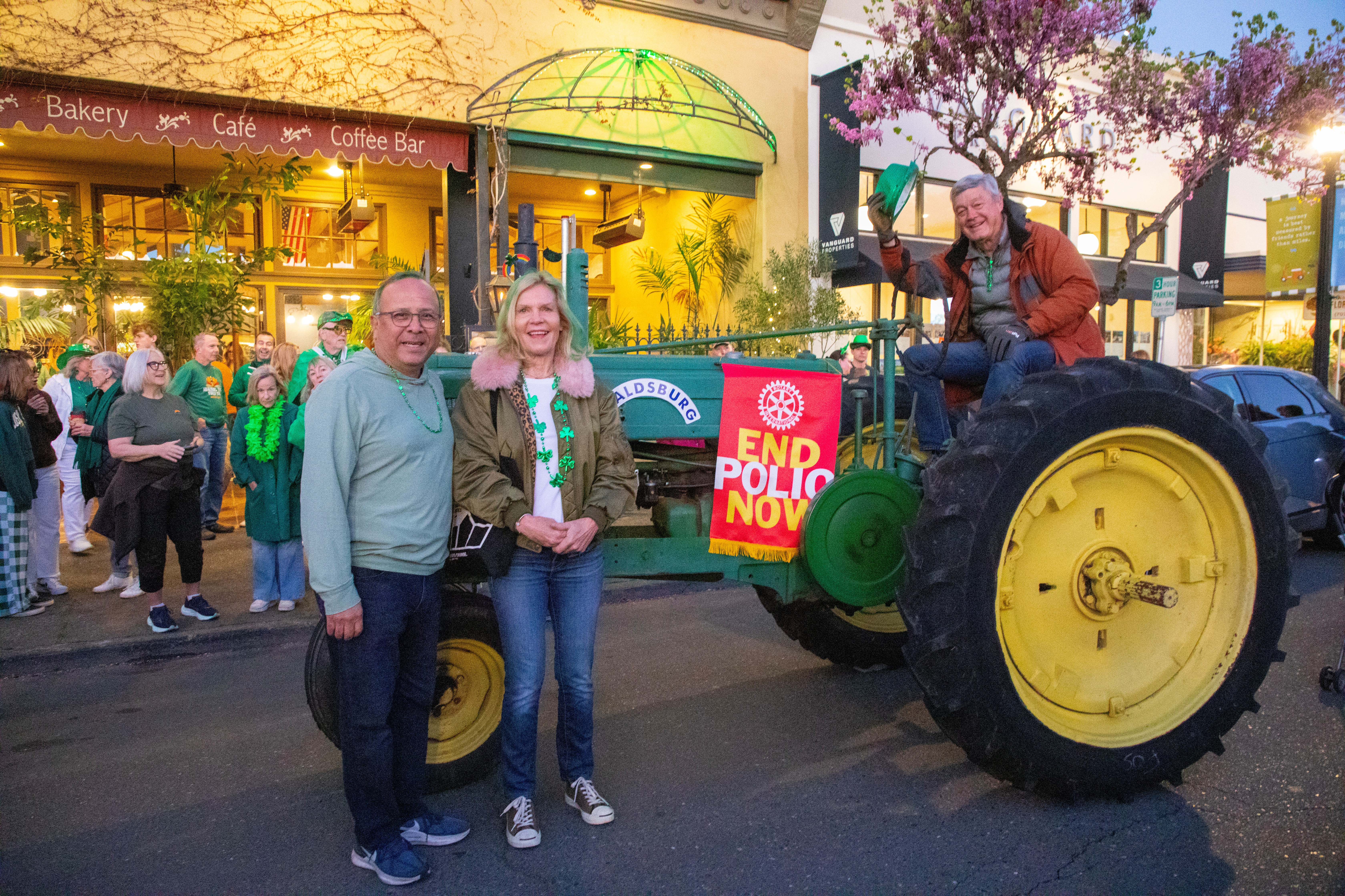 Two people standing beside a vintage tractor at an outdoor event, with a Rotary sign and gathered attendees nearby.