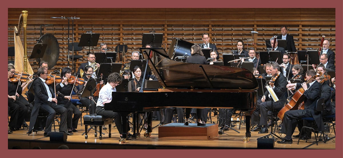 Pianist performing at a grand piano with an orchestra seated on stage in a concert hall.