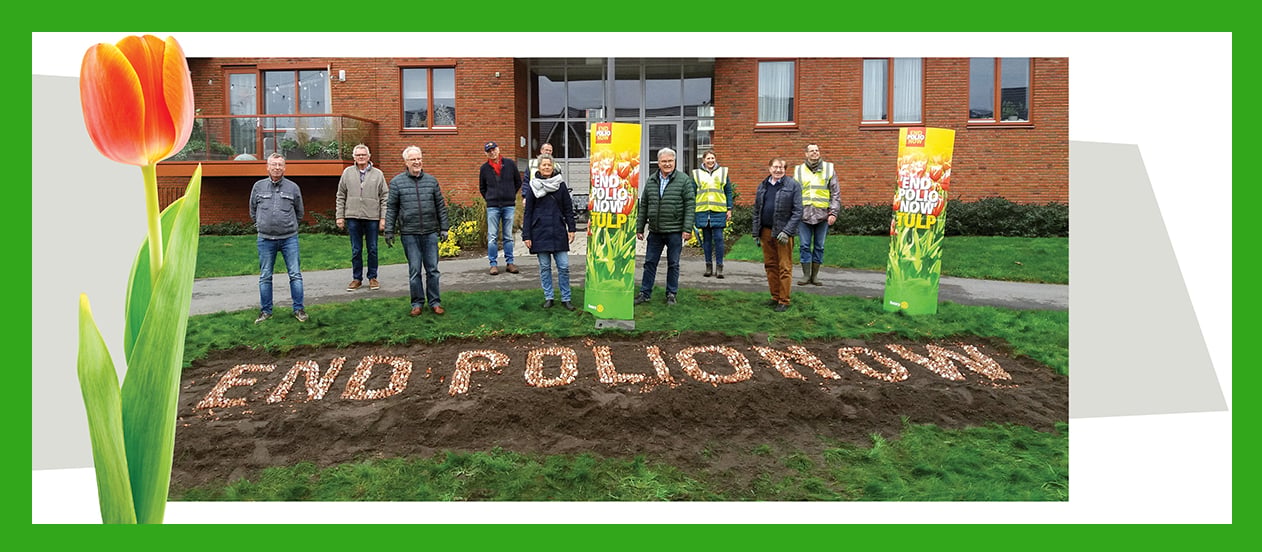 People standing behind a flower bed displaying the text "END POLIO NOW" outside a brick building.