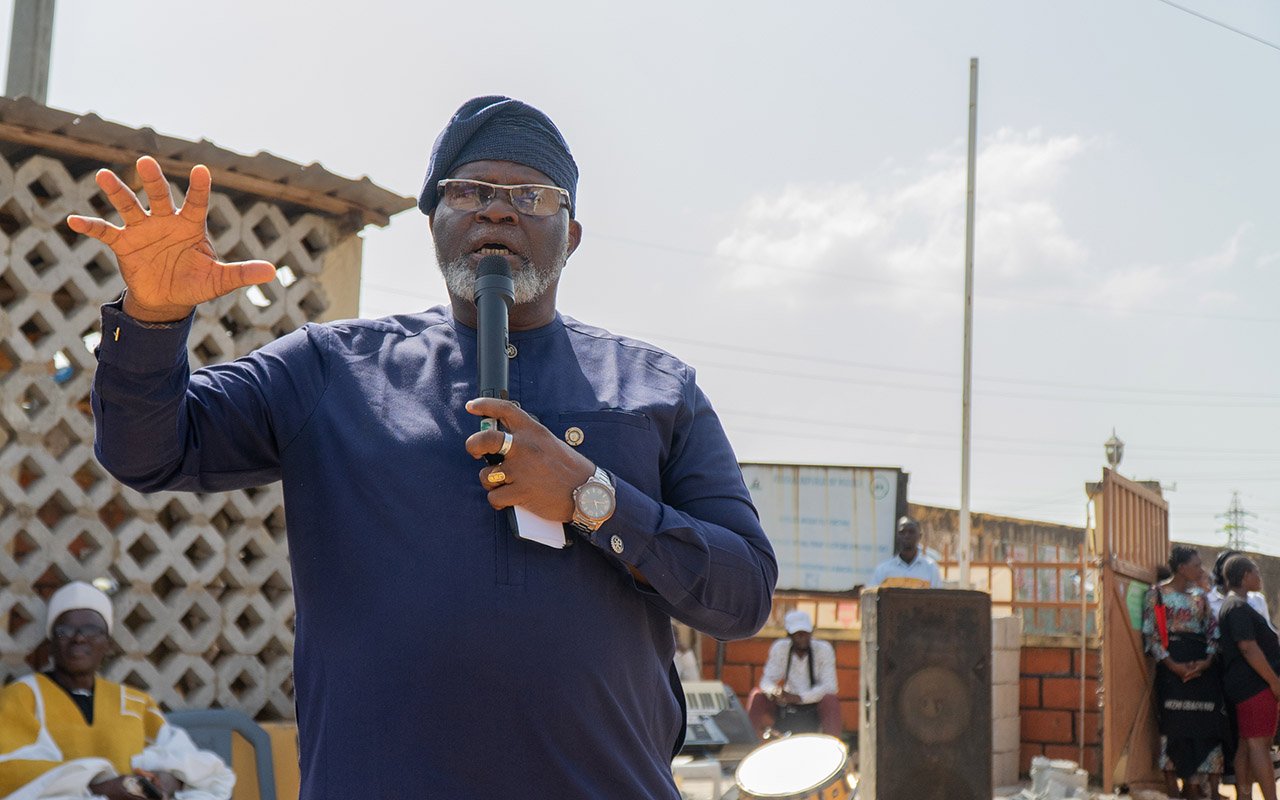 Emmanuel Adedolapo Lufadeju speaking into a microphone and gesturing with an outstretched hand at an outdoor gathering.