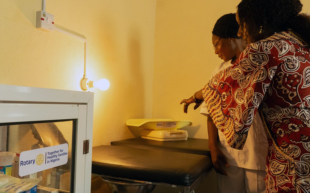 Charity James (left) and Ashezi David Alu Person pointing to a newborn lying on a scale while another person observes the measurement.