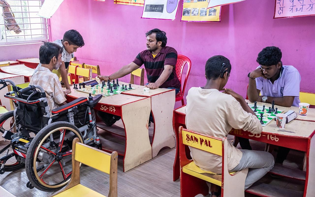 People seated at classroom desks playing chess, including a participant using a wheelchair, during a chess activity.