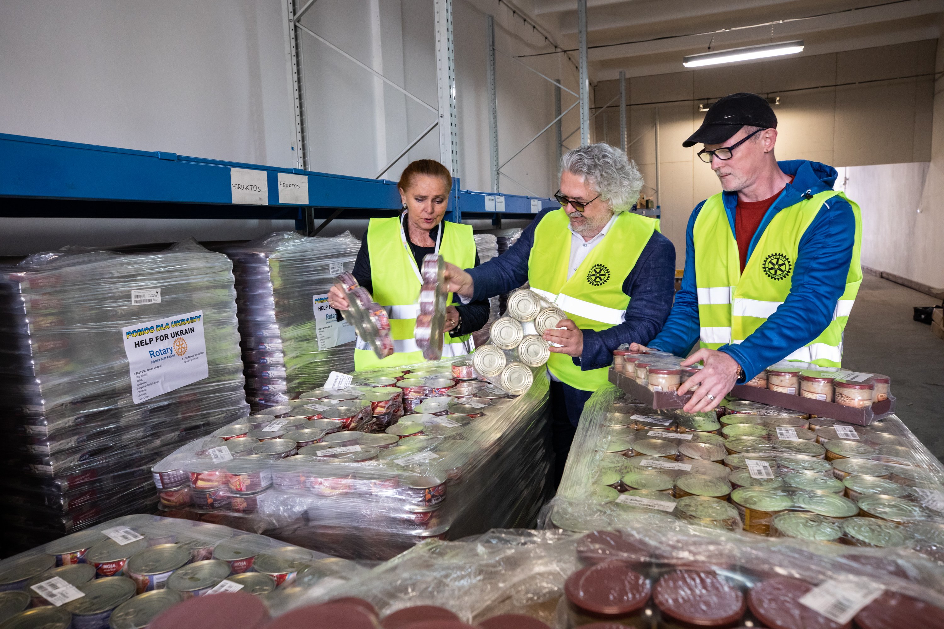 Três voluntários, vestindo coletes de alta visibilidade e toucas, separam alimentos embalados em um ambiente de armazém.
