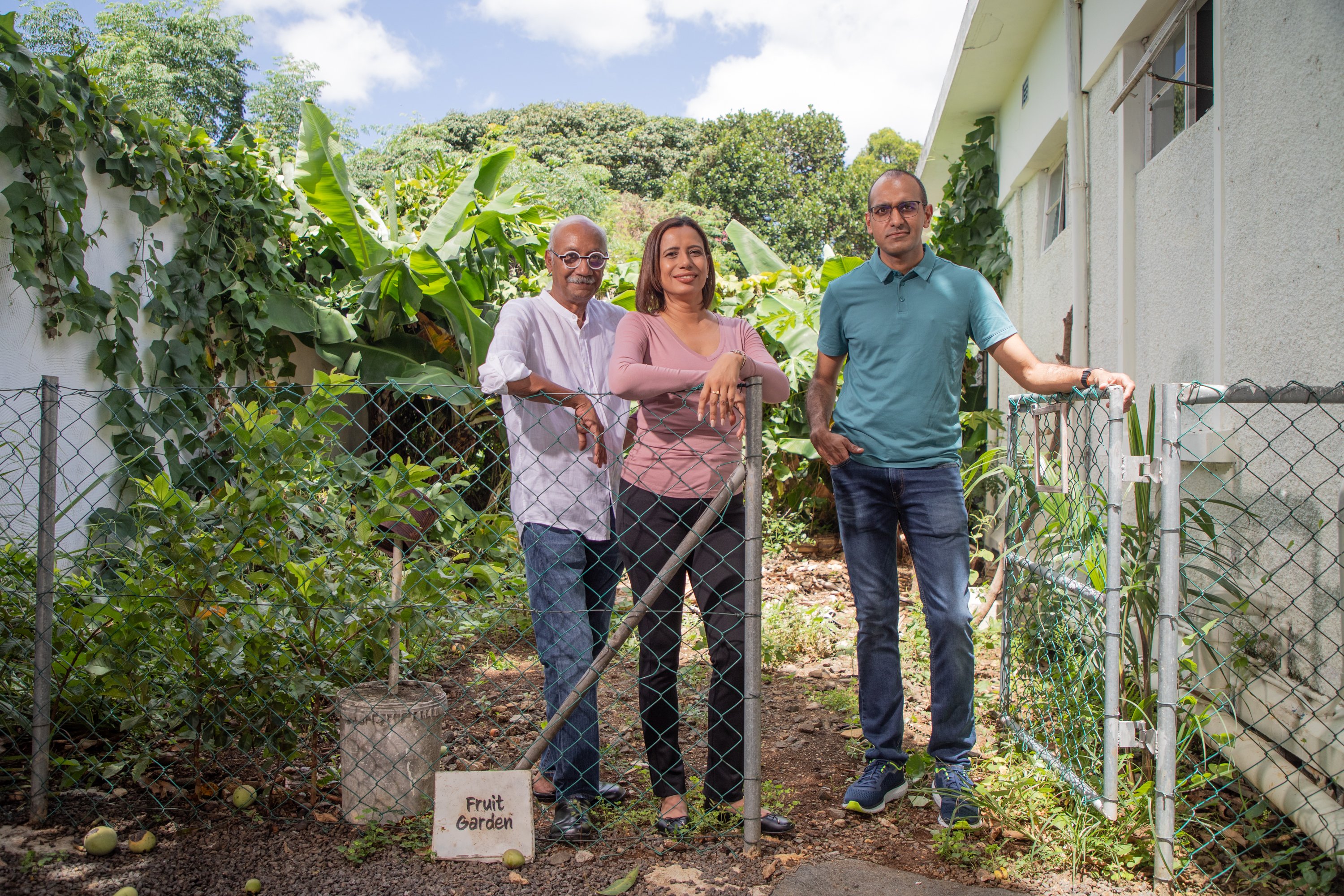 Três pessoas estão em um jardim, segurando ferramentas de jardinagem, perto de canteiros elevados e da parede de um prédio.