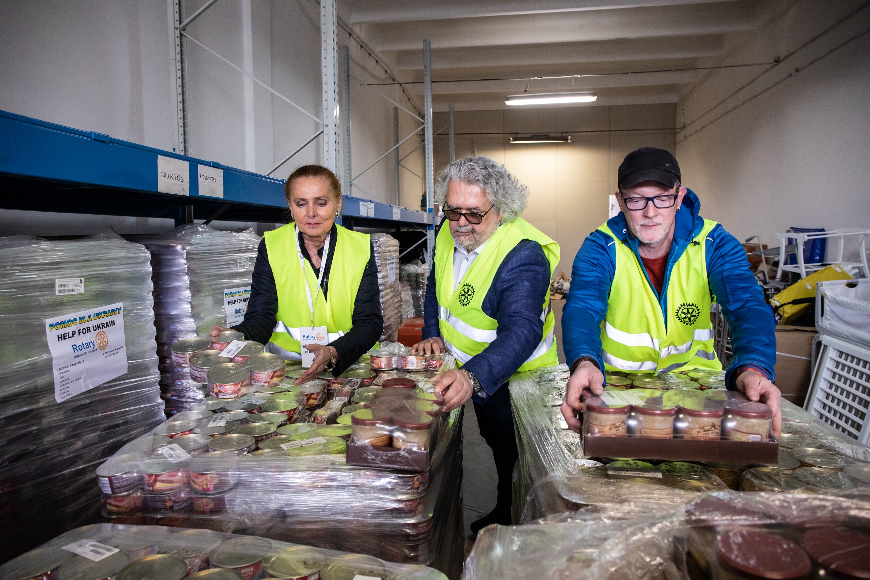 Volunteers in safety vests sort and move boxes of supplies on carts inside a warehouse.