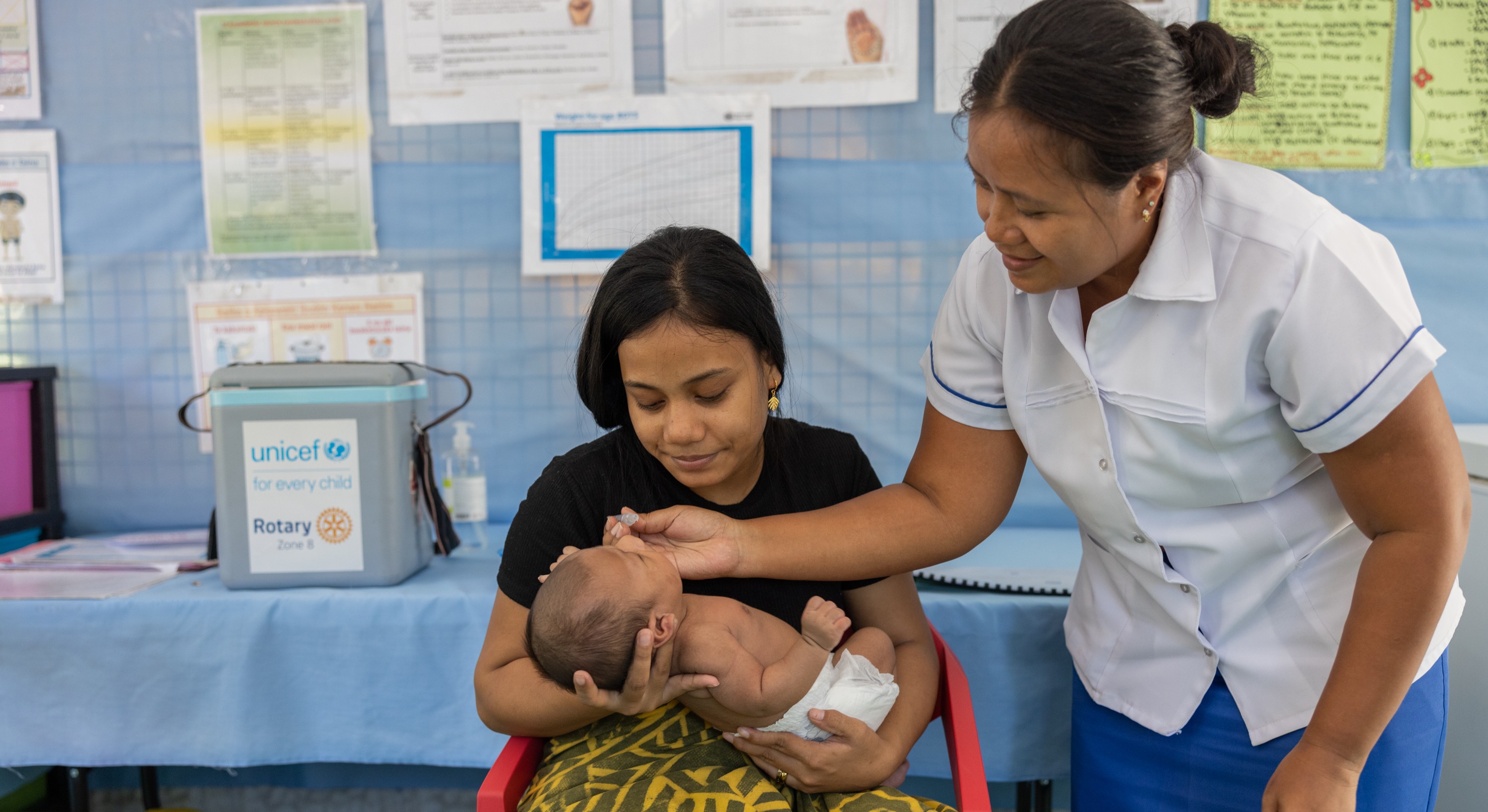 Healthcare worker administers oral vaccine drops to an infant held by an adult in a clinic.