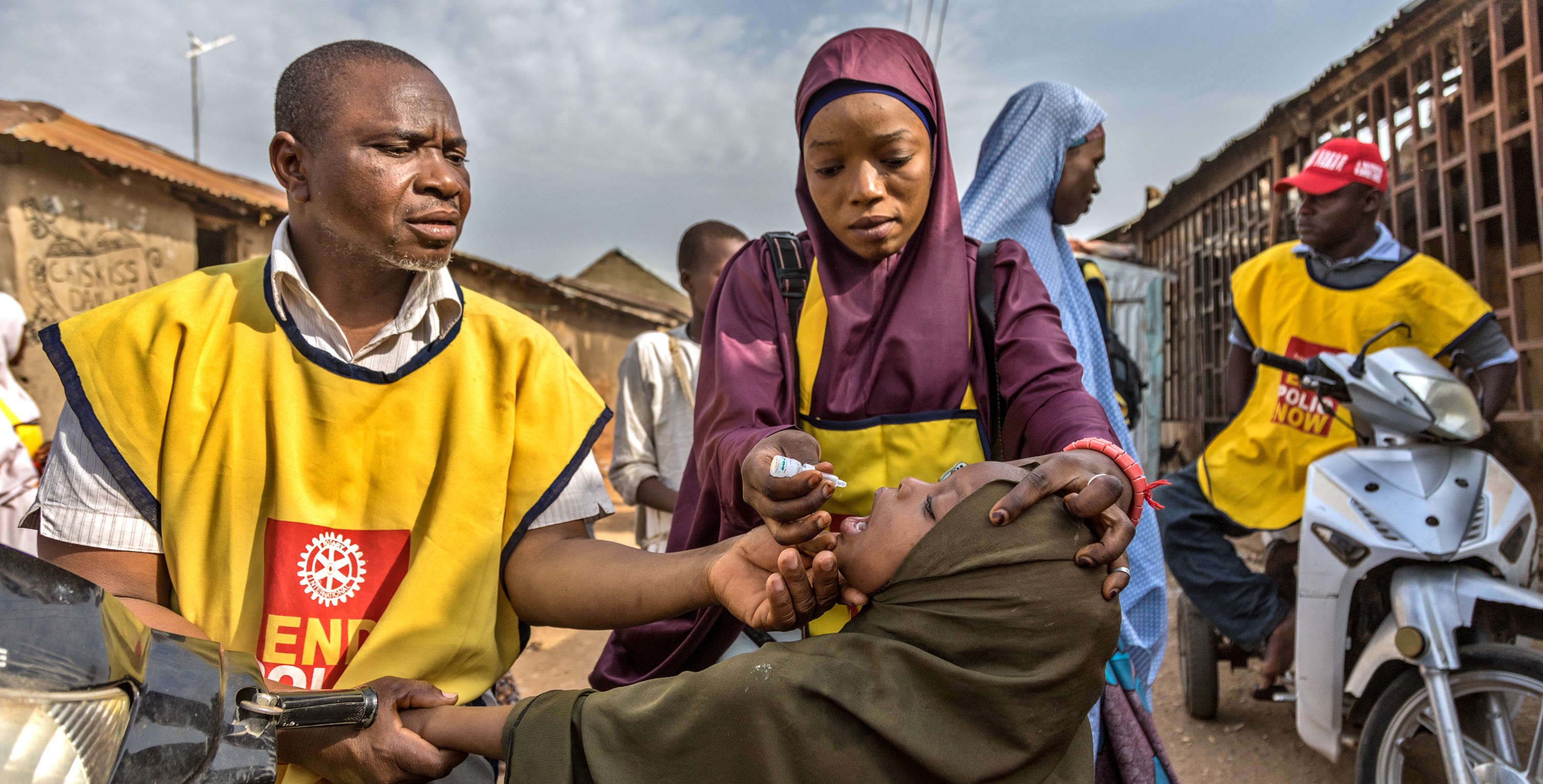 Health workers in yellow “End Polio Now” vests give oral polio drops to a child during a door-to-door campaign.