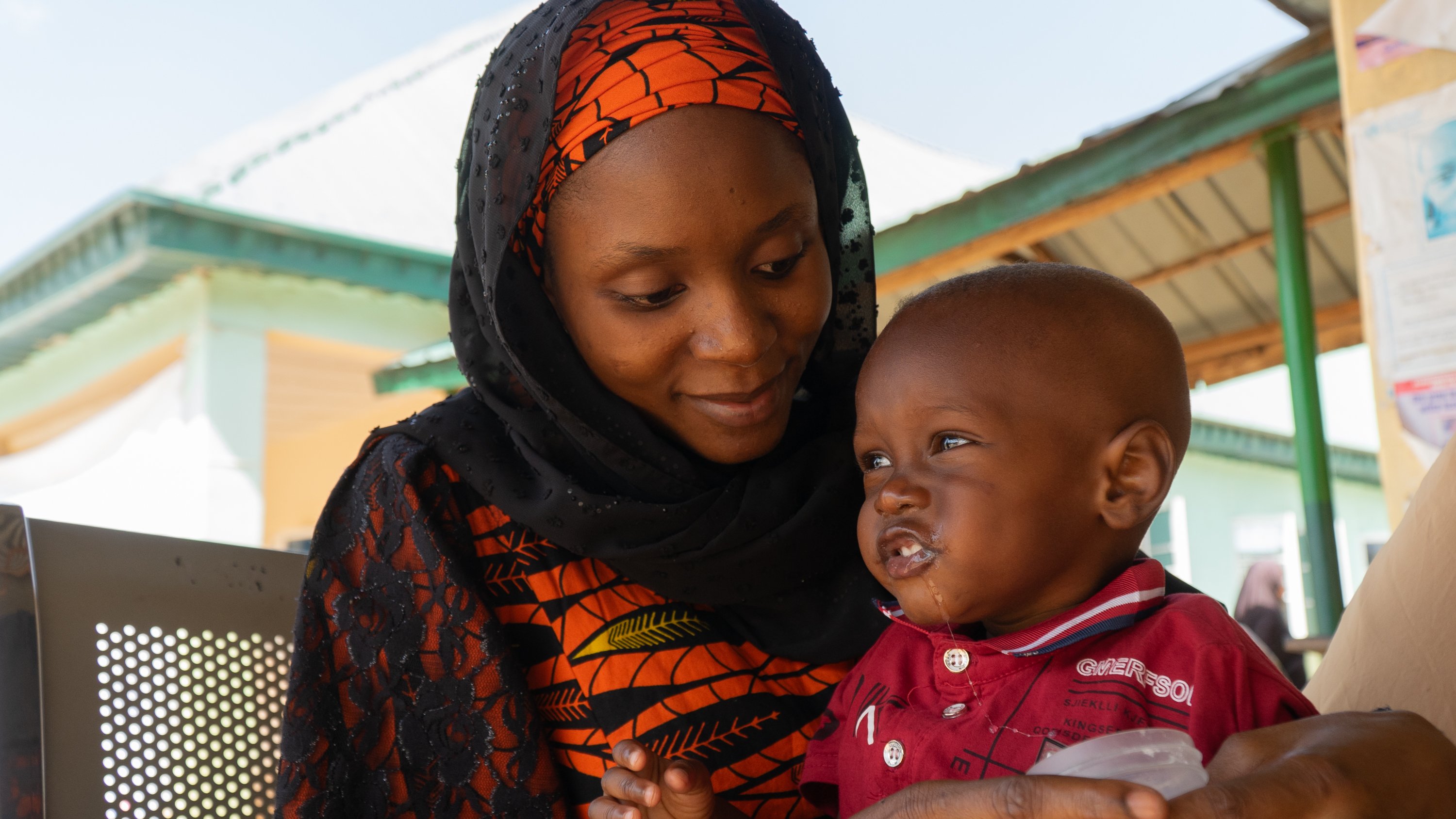Adult holds a child while waiting for care at a healthcare facility, with clinic buildings visible in the background.