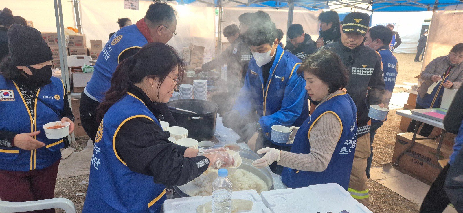 Volunteers in vests prepare and serve food from steaming pots at a temporary outdoor relief station.