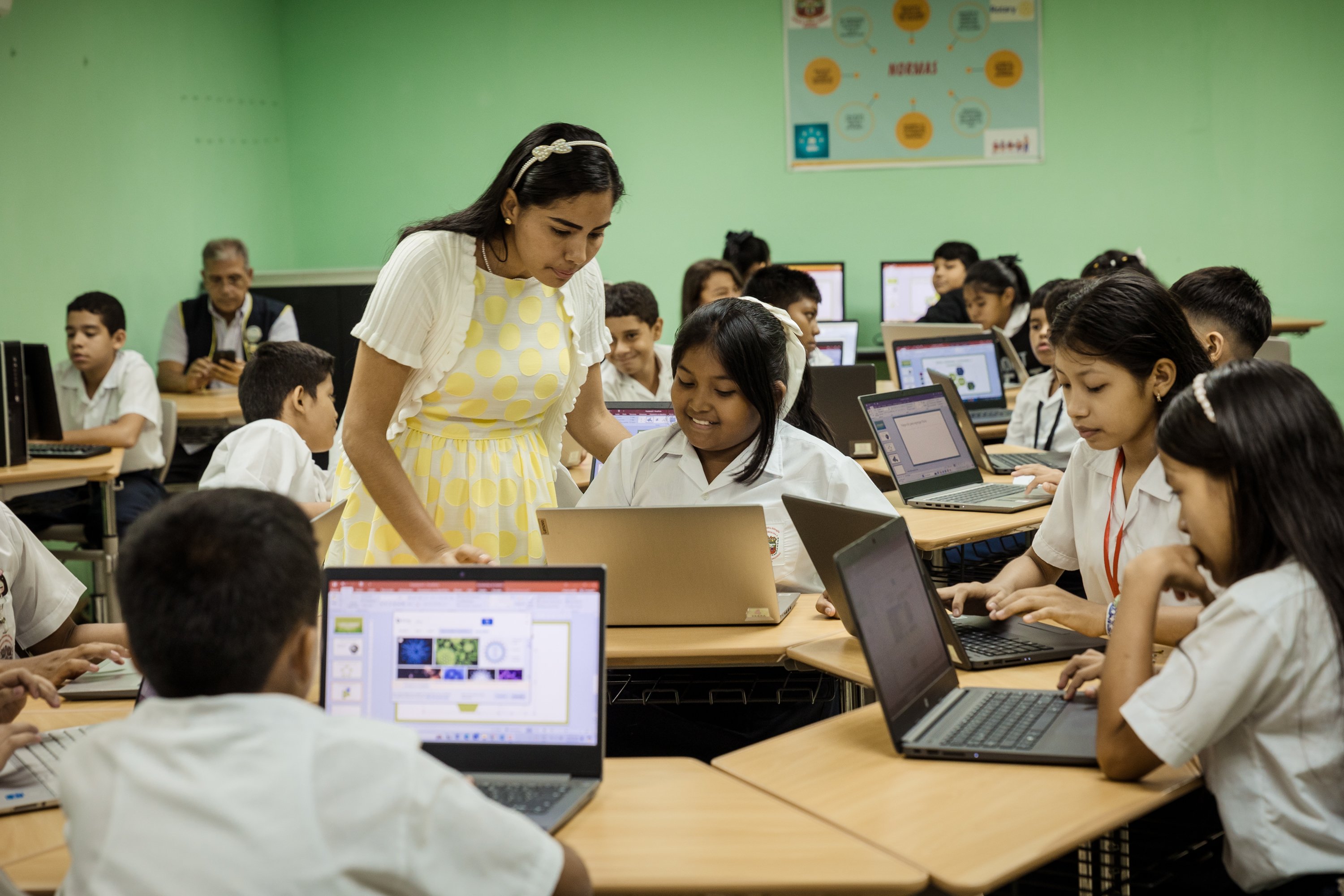 Instructor assists students using laptops at desks in a classroom with digital learning equipment.