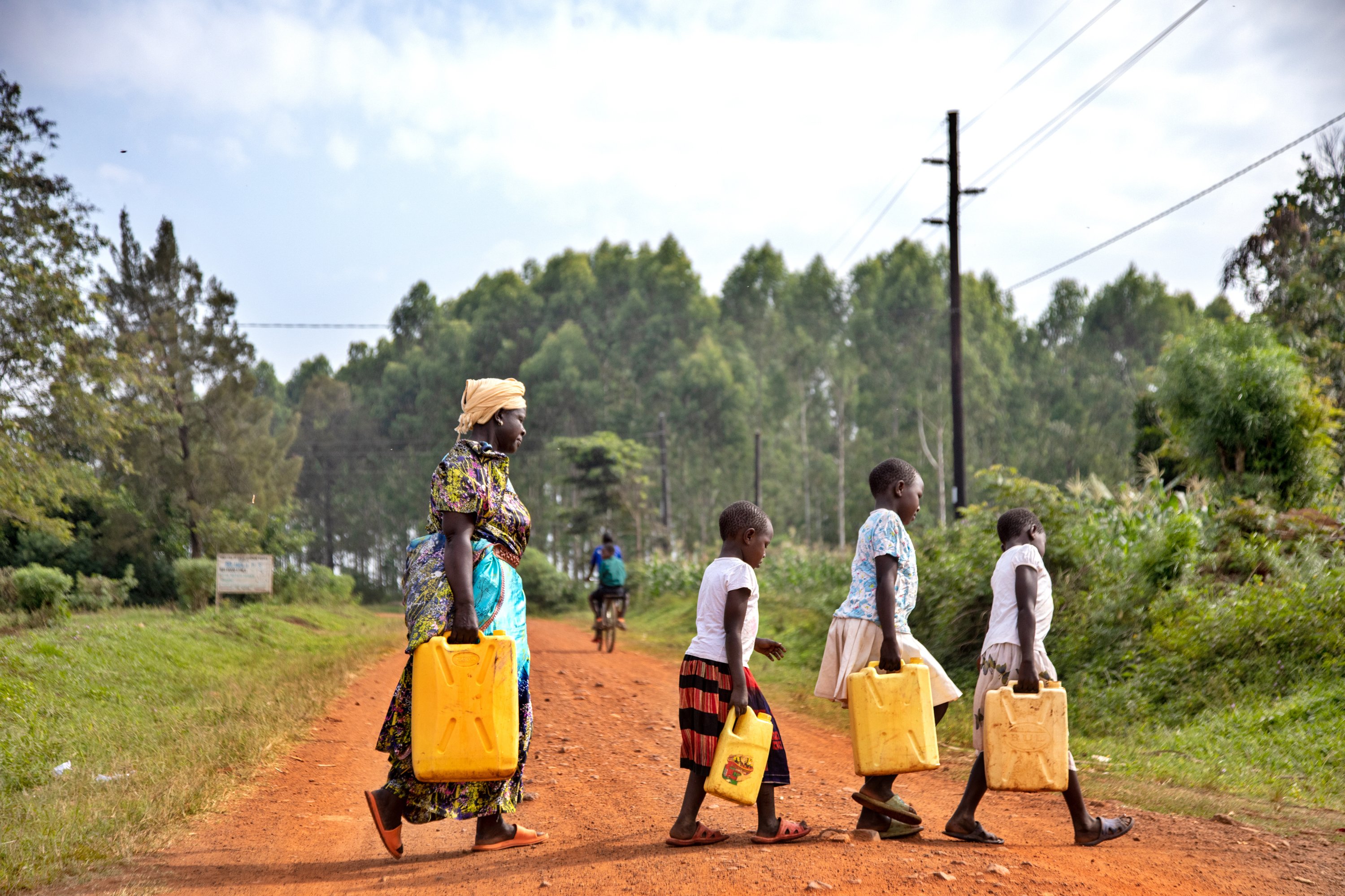 People carry yellow jerrycans of water along a dirt road near a borehole well in a rural area.