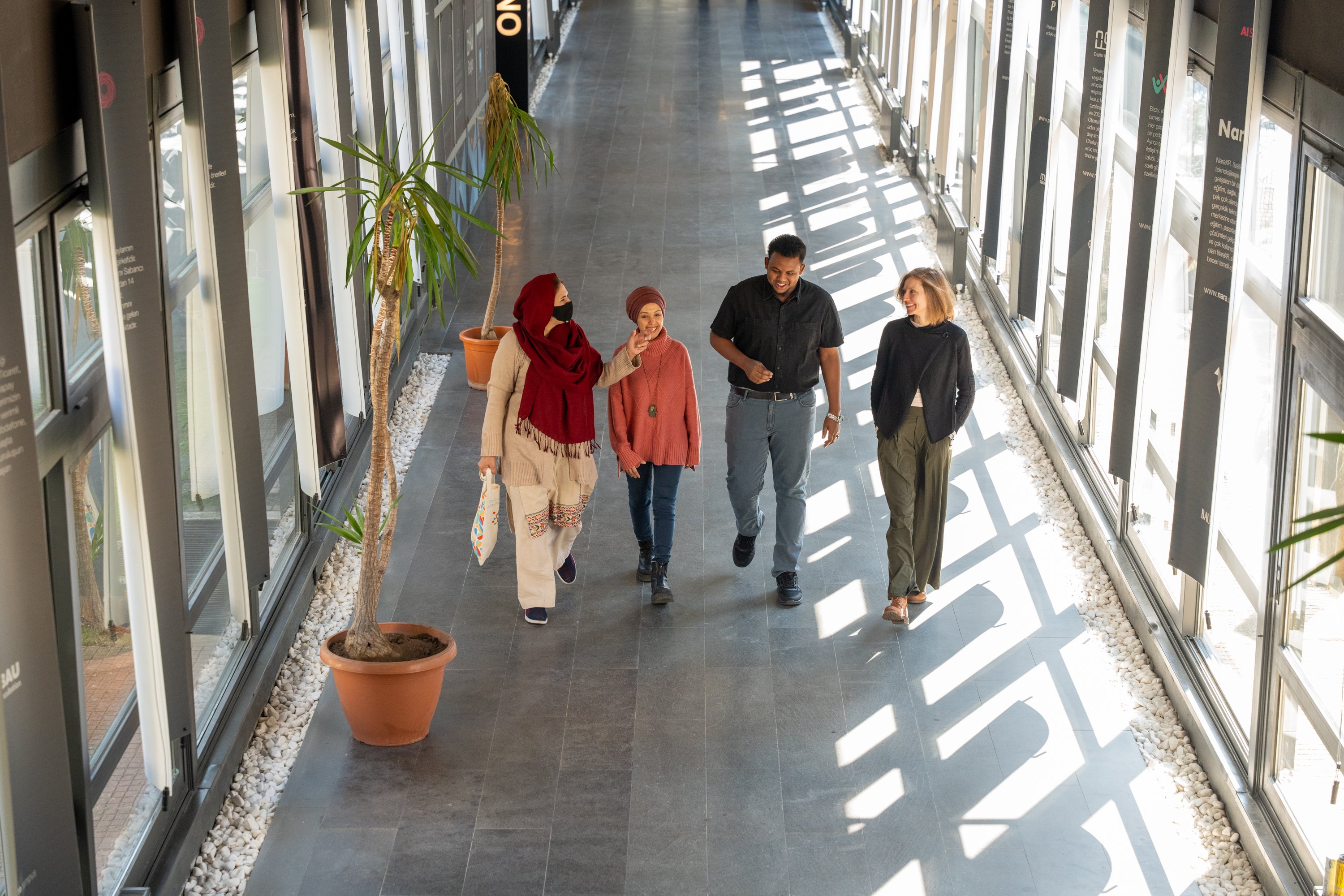 Four people walk down a sunlit corridor with large windows on both sides inside a modern building.