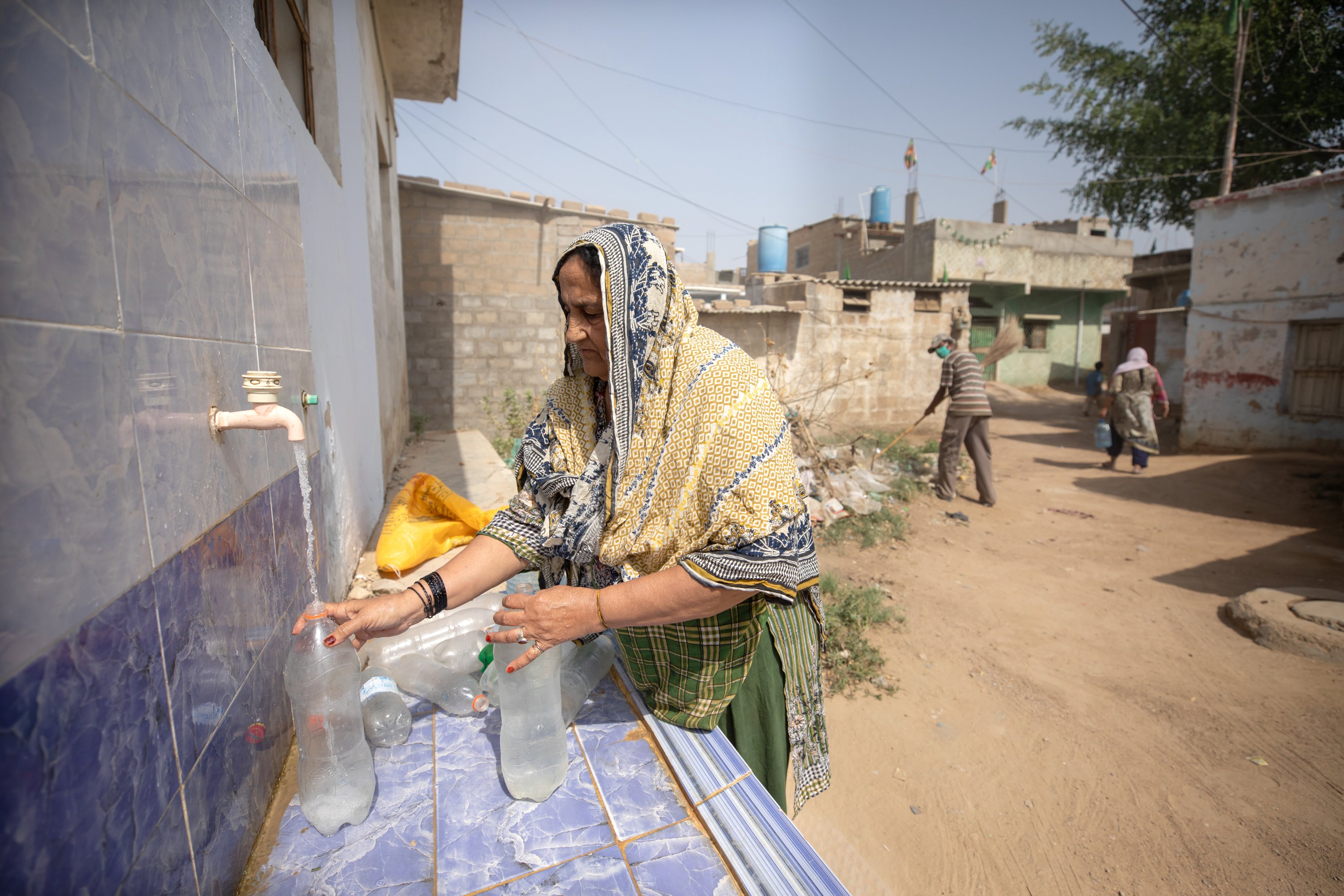 A woman fills a container from a water tap at an outdoor filtration station along a street with nearby buildings.