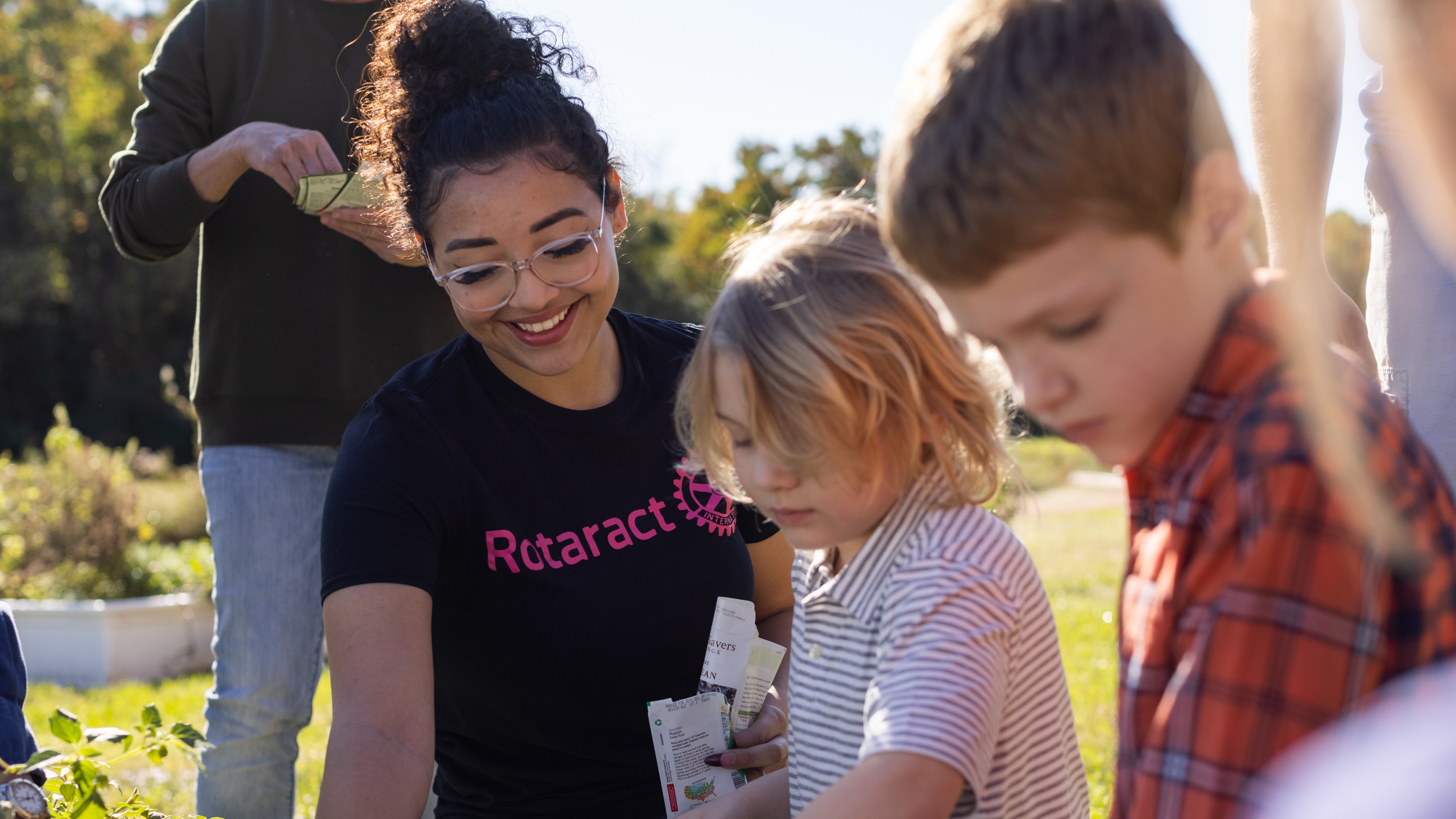 Un adulto con una maglietta con la scritta “Rotaract” aiuta due bambini a piantare qualcosa nel terreno all’aperto usando degli attrezzi da giardinaggio.