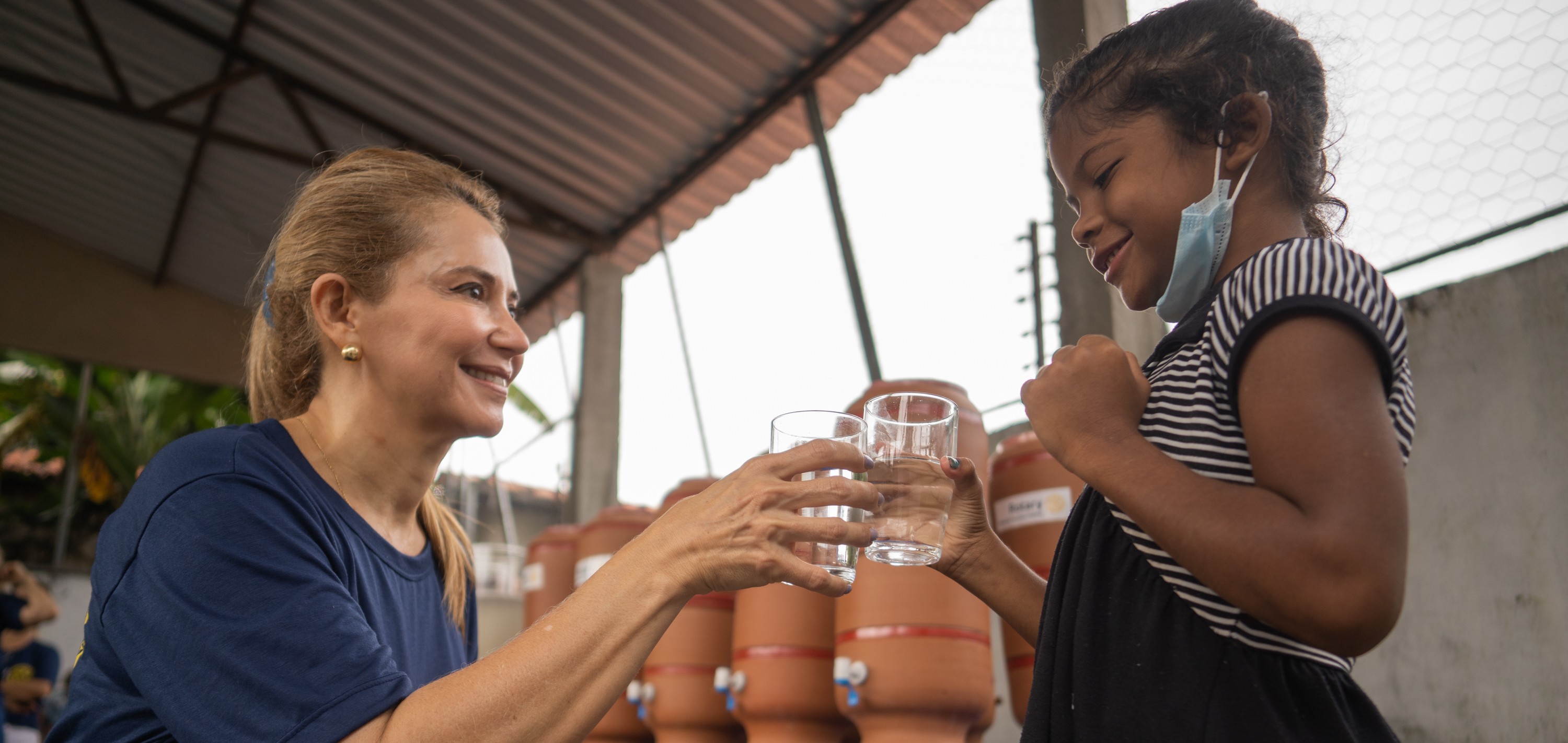 An adult and a child hold glasses of filtered water poured from a clay container at an outdoor table.