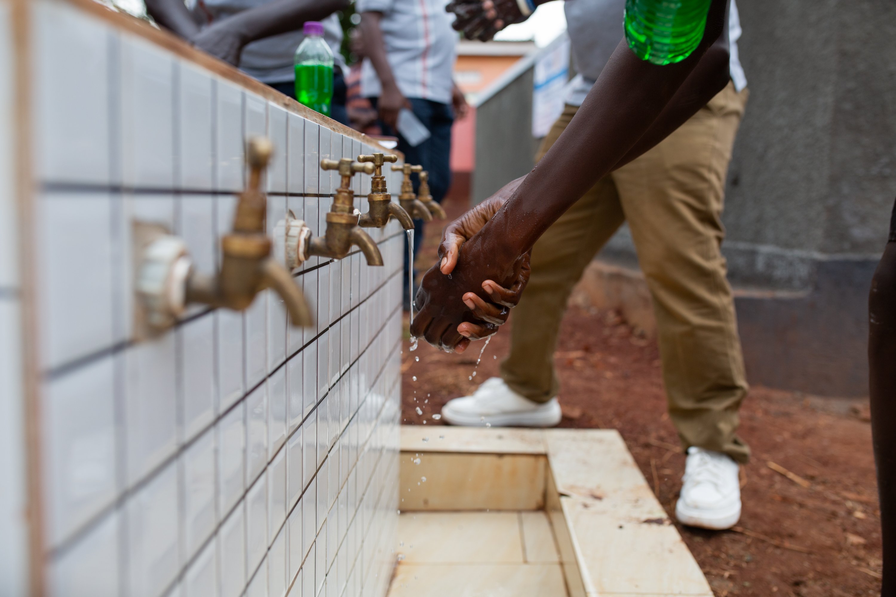 Person washes hands under multiple outdoor taps connected to a tiled water station.