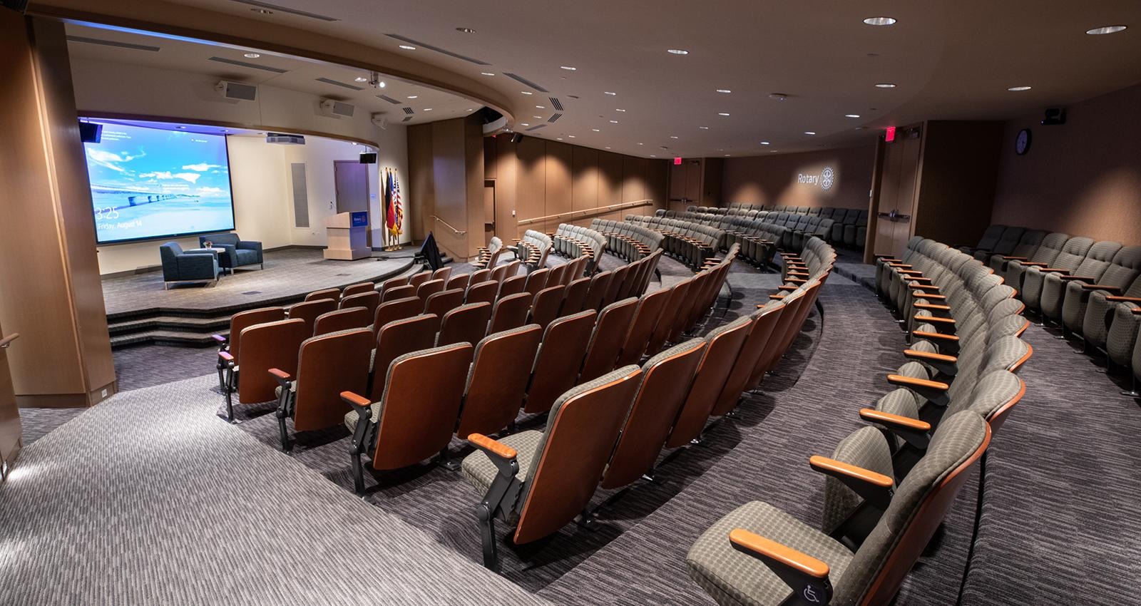 Curved rows of auditorium seating face a large presentation screen and stage in a conference hall.
