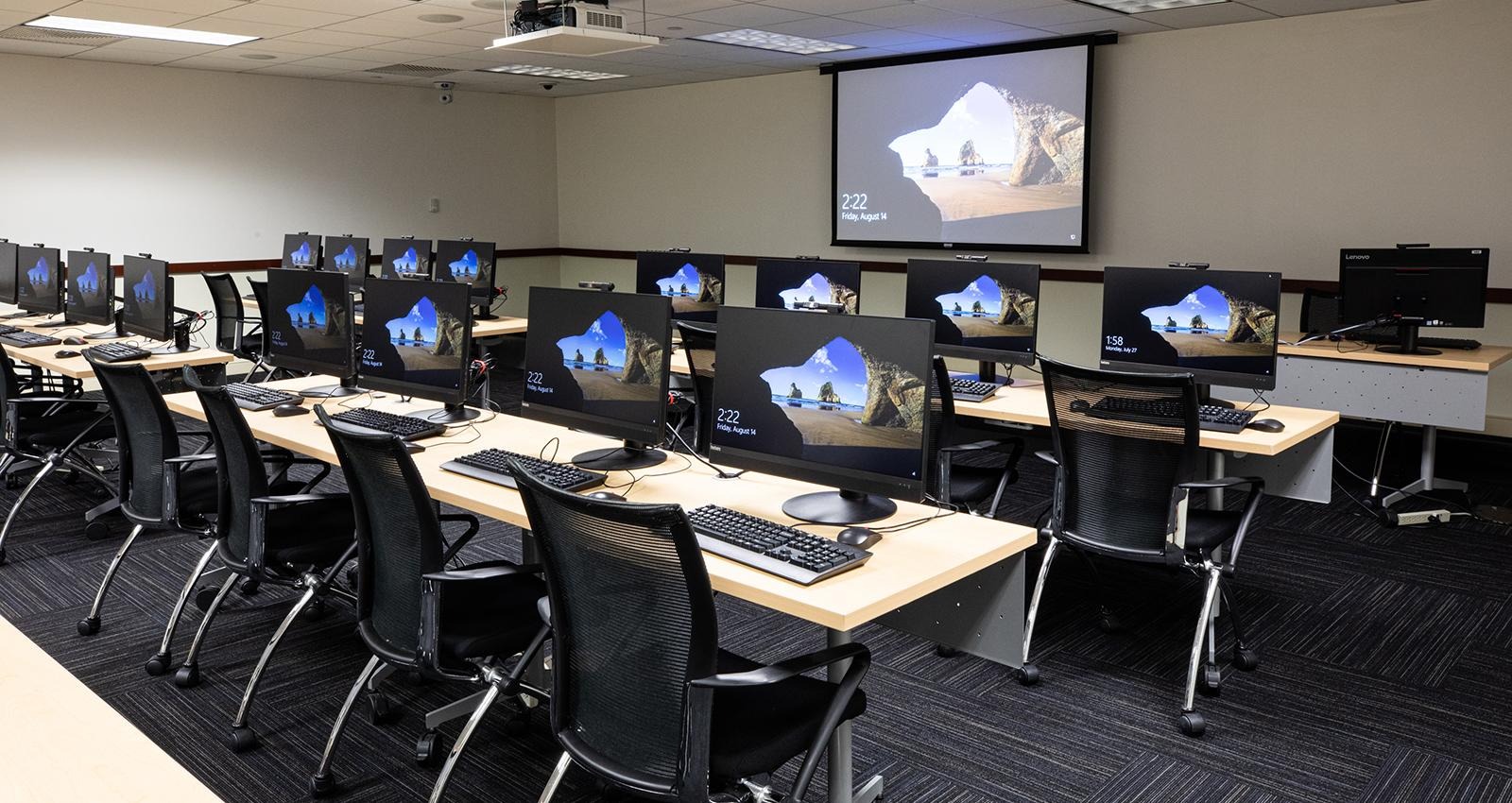 Rows of desks with multiple desktop monitors displaying the same screen in a computer lab classroom.
