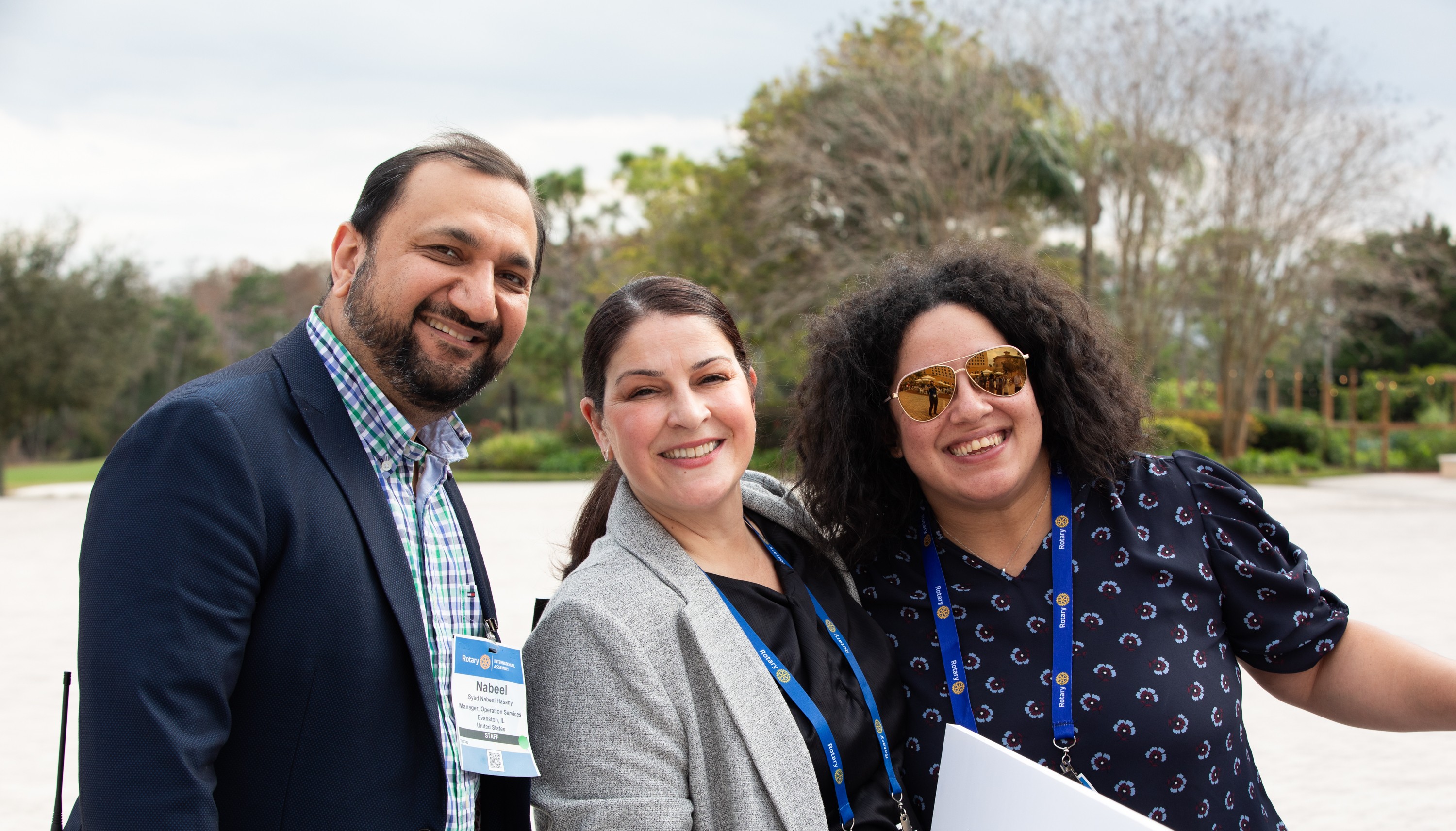 Three people wearing lanyards stand outdoors holding a white geometric sign together.