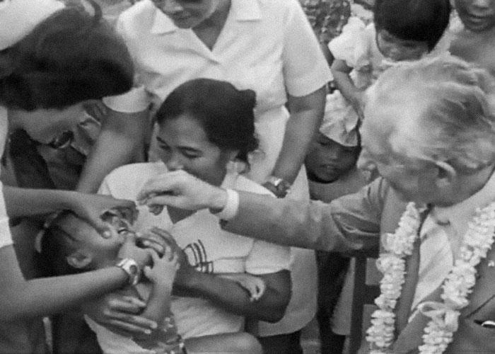 Adult administers oral drops to a child while others assist and observe in a crowded setting.