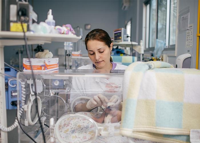 Um profissional de saúde acomoda um recém-nascido dentro de uma incubadora, em meio a equipamentos médicos, em uma unidade de terapia intensiva neonatal.