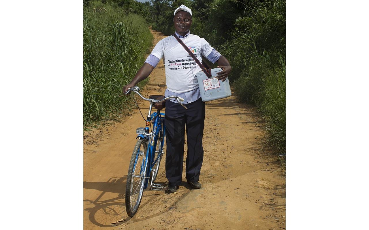 Um profissional de saúde está ao lado de uma bicicleta, segurando uma sacola e materiais, em um caminho de terra ao ar livre.