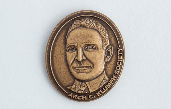 Alfinete de lapela de bronze com retrato em relevo; a inscrição “Sociedade Arch Klumph” aparece ao redor da borda.
