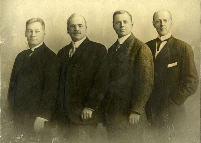 Quatro pessoas vestidas de terno estão lado a lado em um retrato formal de estúdio, contra um fundo liso.