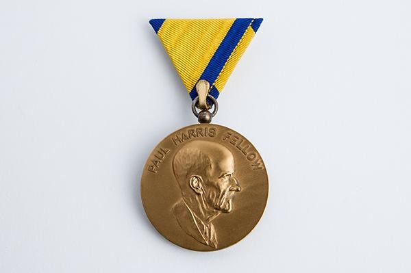 横顔の肖像が刻まれた丸い金メダルで、上部には青と黄色の三角形のリボンピンが付いています。