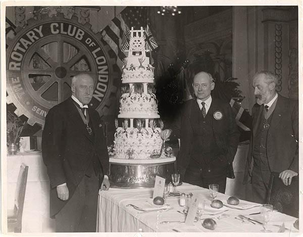 Trois personnes se tiennent debout à côté d'un gâteau à étages, assises à une table, avec l'emblème du « Rotary club » affiché sur une toile de fond derrière elles.