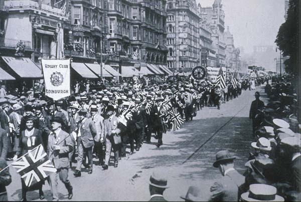 Une foule défile dans une rue de la ville en brandissant des drapeaux et des banderoles, tandis que les passants se pressent sur les trottoirs.