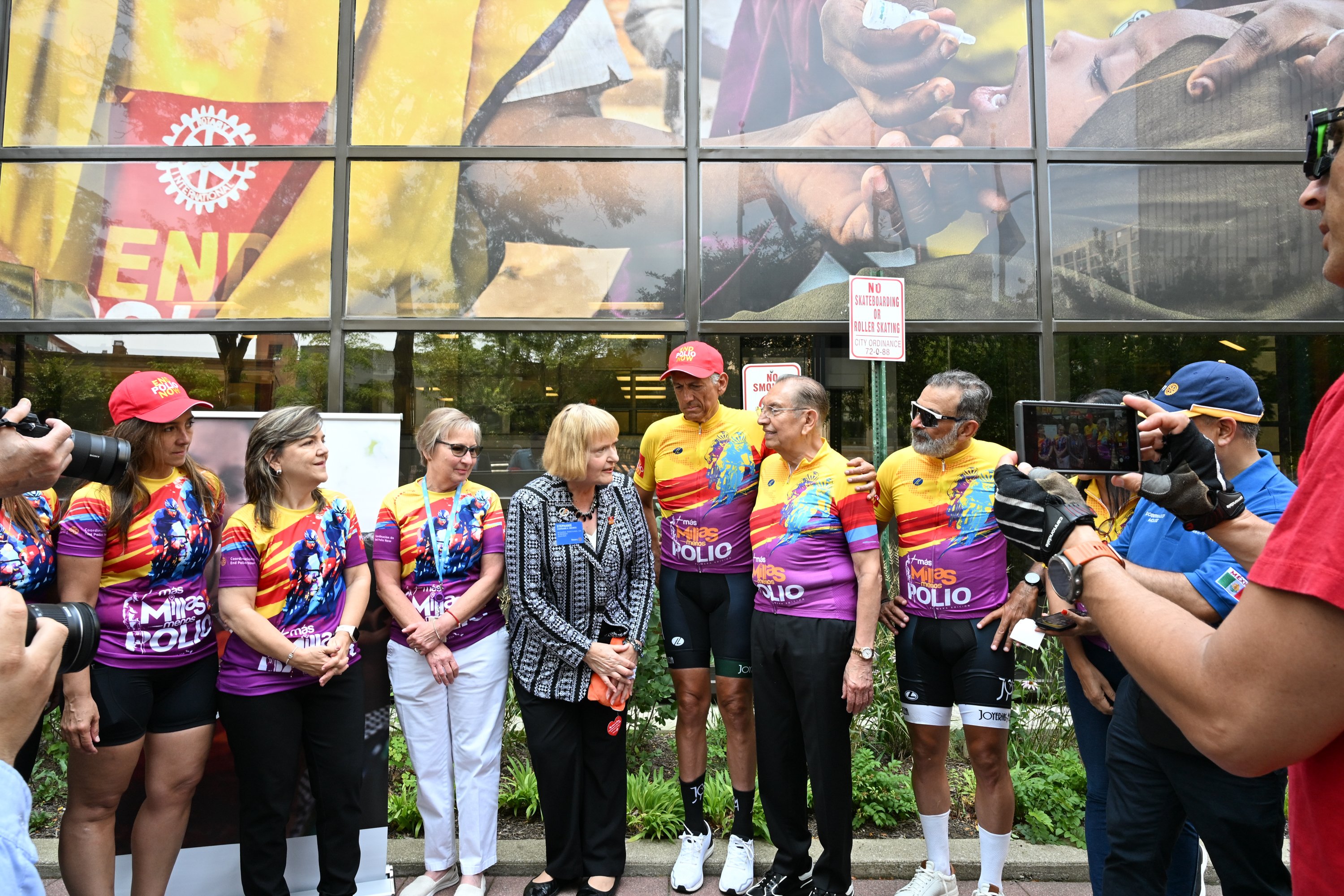 Radfahrer in bunten Trikots stehen vor dem Hauptsitz von Rotary International, während Fotografen in der Nähe des „Rotary International“-Schilds Fotos machen.