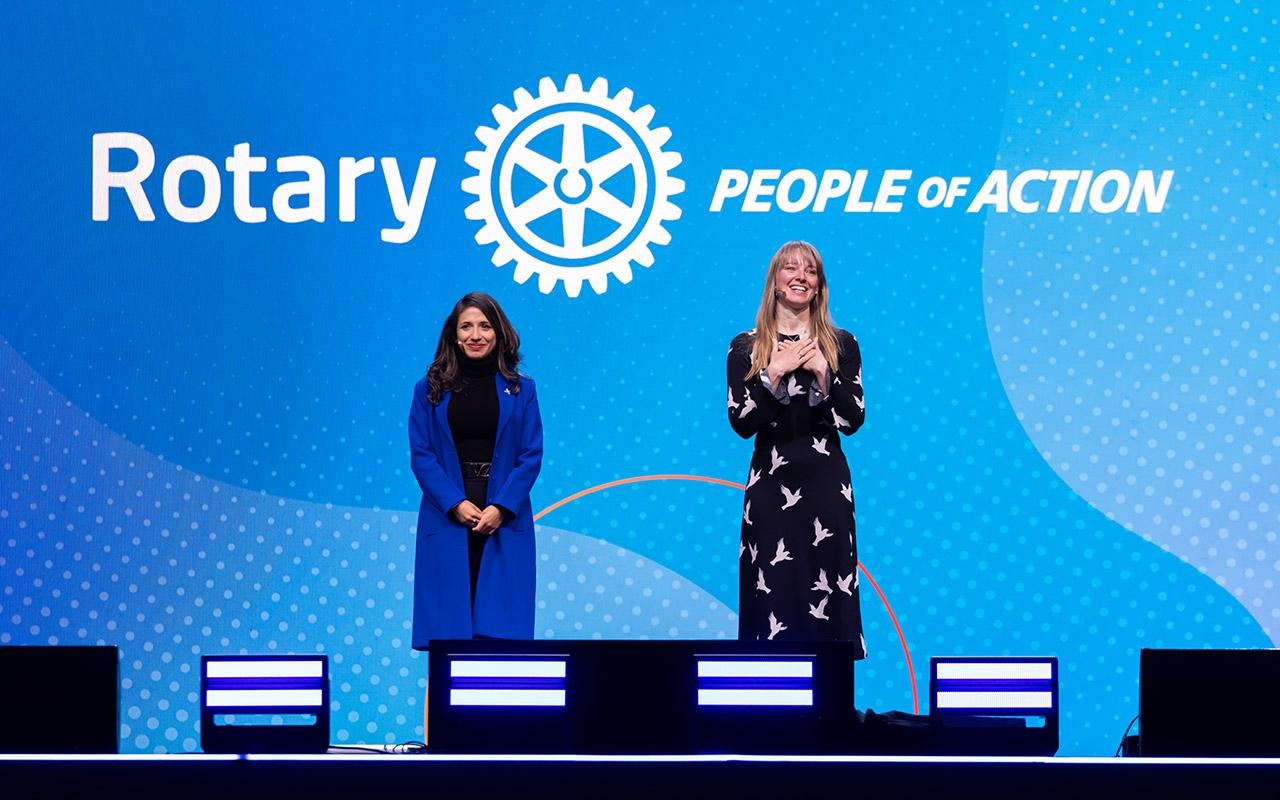 Zwei Personen stehen auf einer Bühne und sprechen vor einem blauen Hintergrund, auf dem „Rotary People of Action - Wir tun was“ zu lesen ist, in ein Mikrofon.