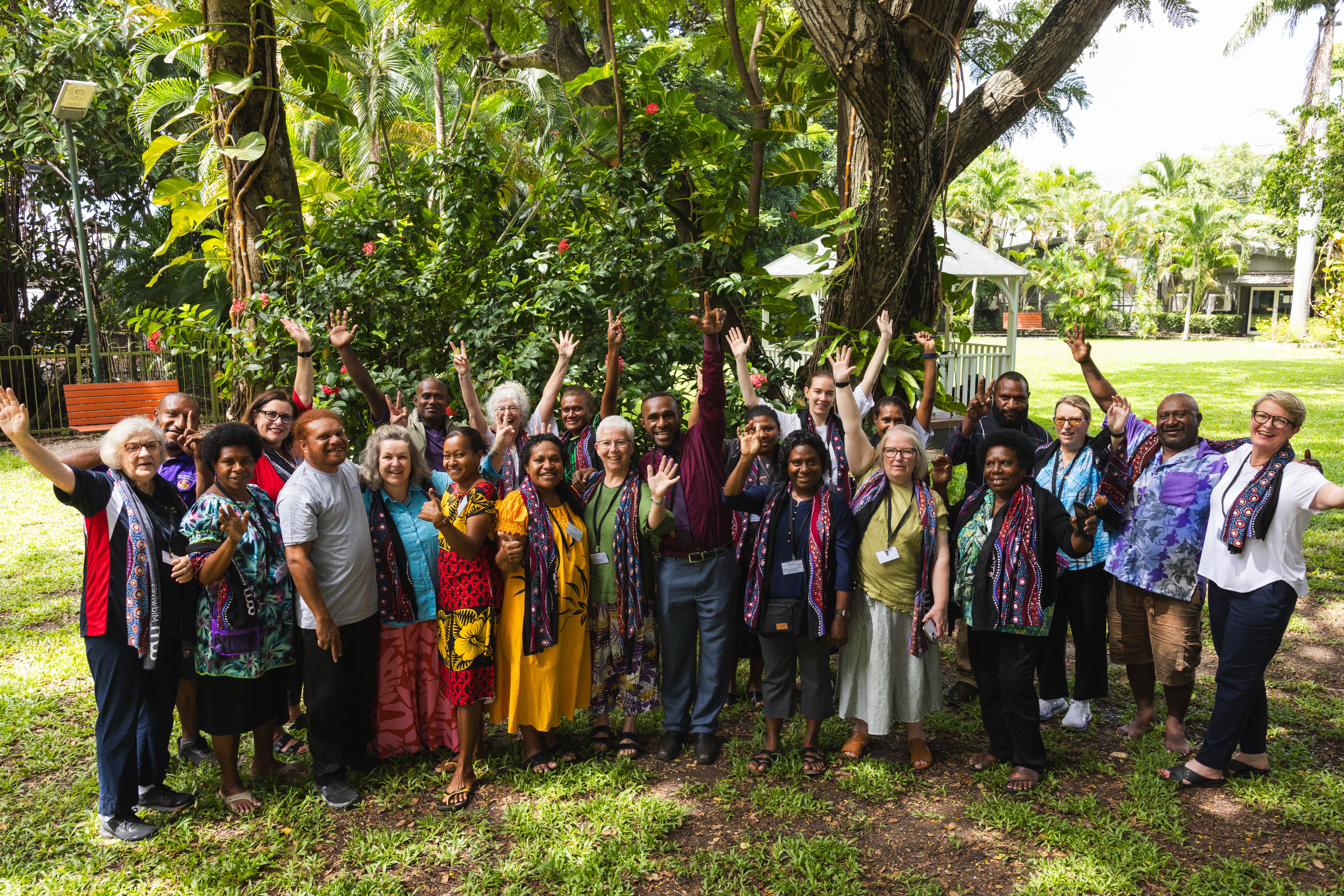 A large group of people stands outdoors under trees, smiling and raising their hands together for a photo.