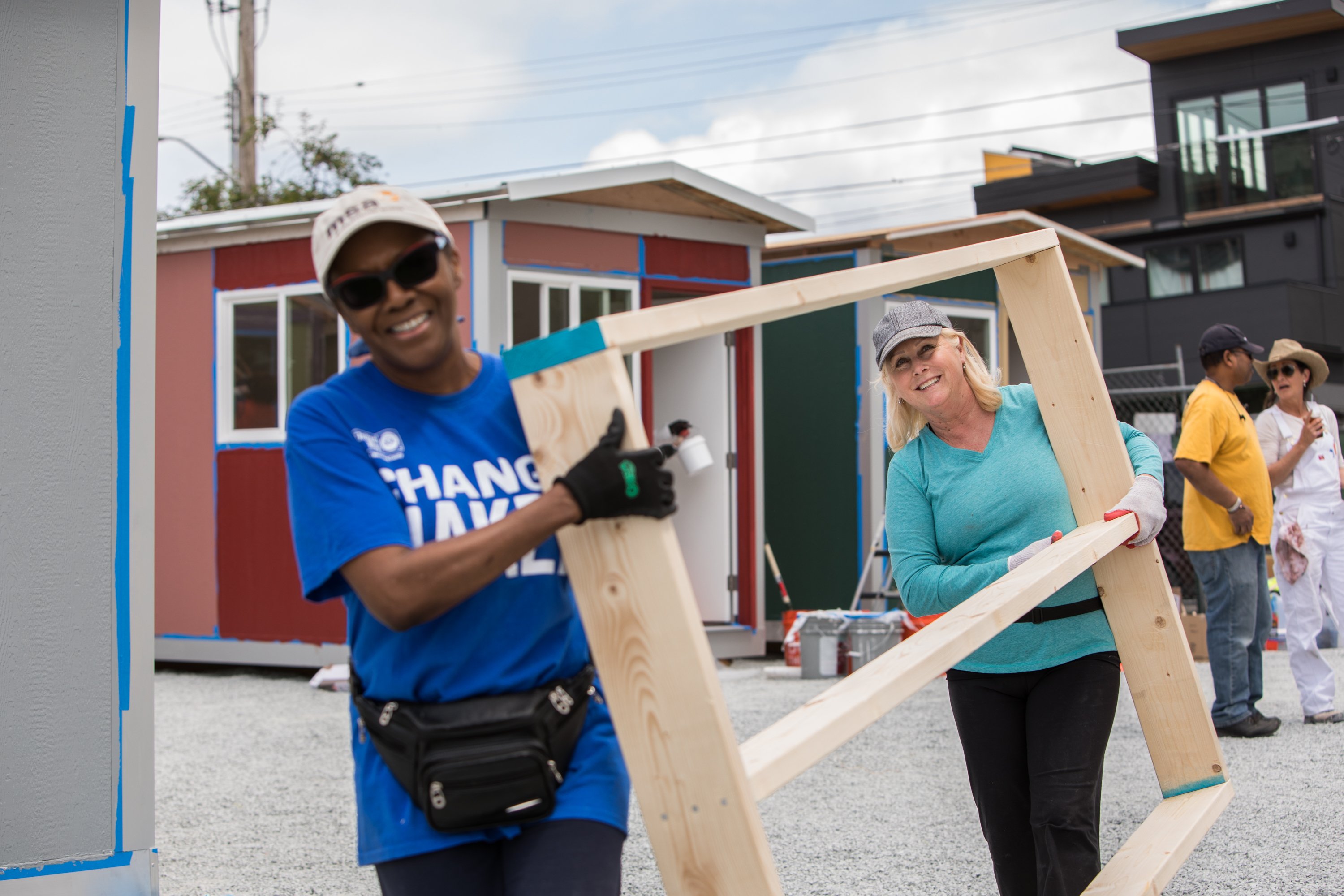 Volunteers carry and position wooden boards while building small painted houses at an outdoor construction site.