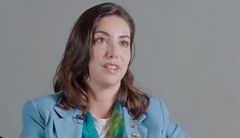 Person speaks during an interview against a plain background with a microphone clipped to clothing.