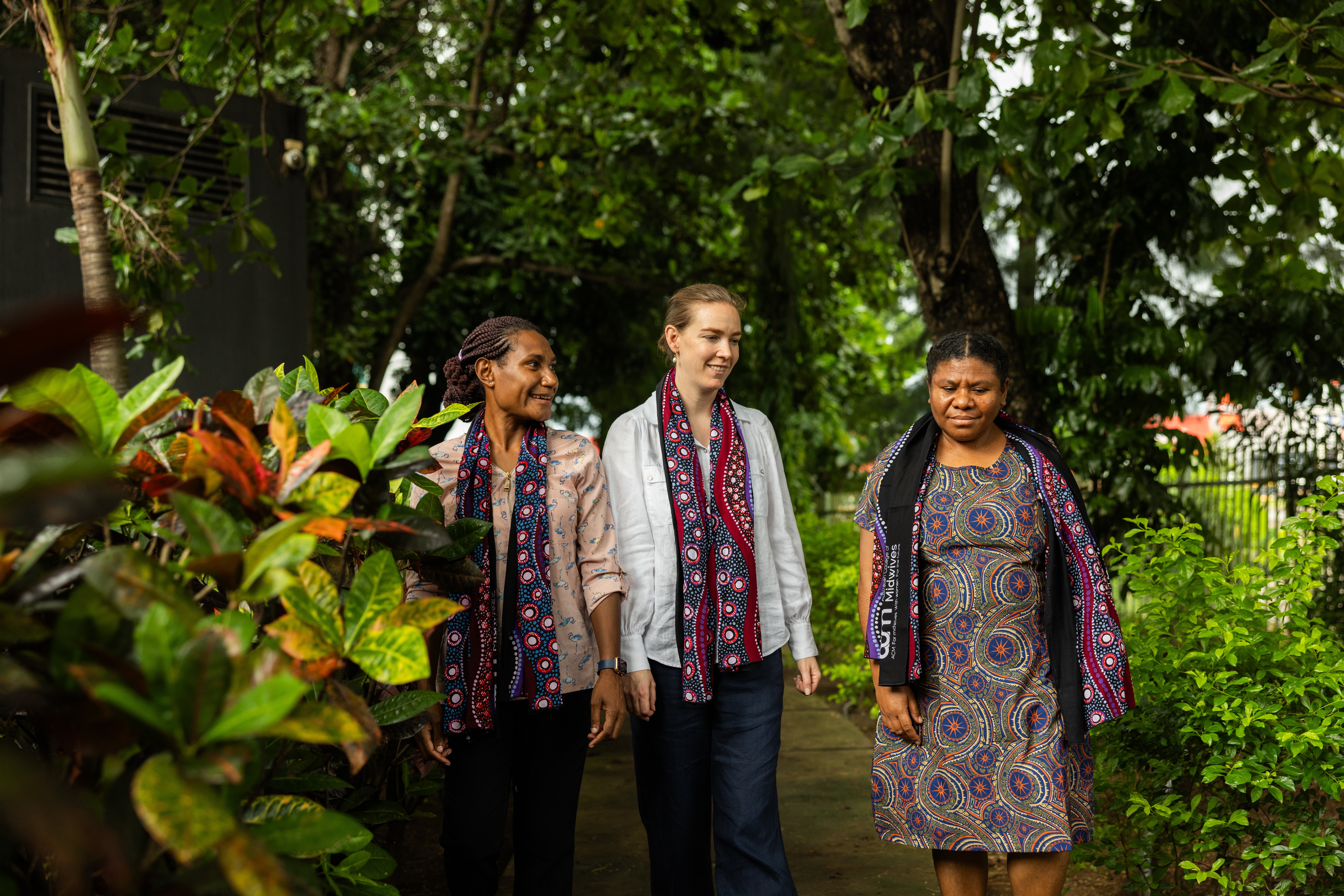 Three people walk along a garden path surrounded by dense greenery and plants.