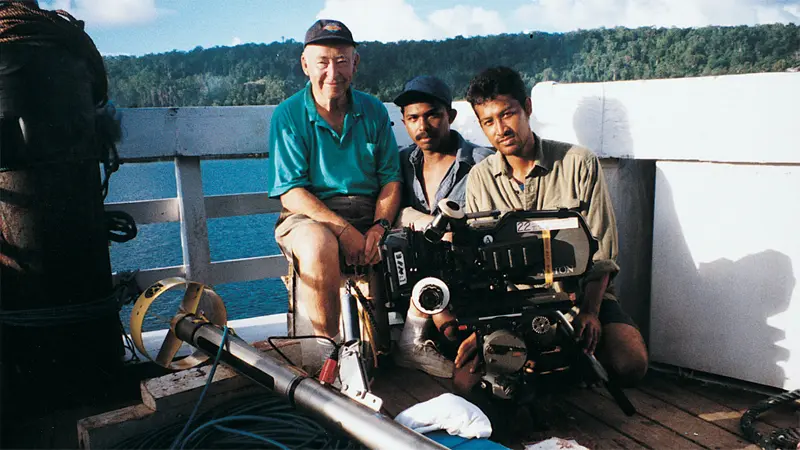 Camera crew sitting on deck, overlooking water and forest.