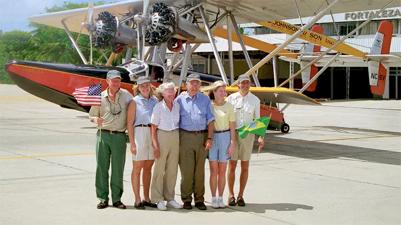 Group poses beside vintage airplane; flags held; airport background.