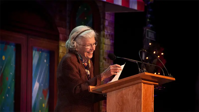 Elderly woman smiling, reading at podium, festive backdrop.