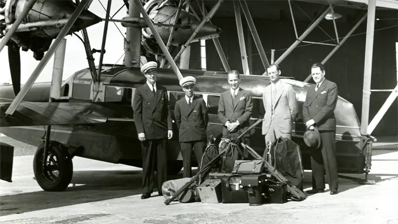 Five men posing beside airplane; luggage on ground. Hangar background.