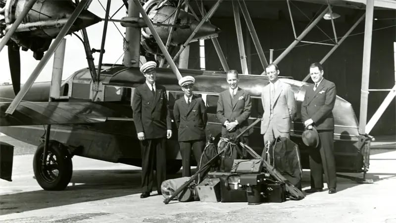 Cinq hommes posant à côté d’un avion ; bagages au sol. Hangar en arrière-plan.